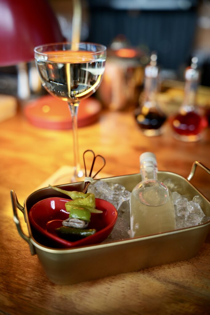 Chilled martini in a coupe beside a brass tray with a gilda skewer in a red heart-shaped dish and a carafe of the remaining pour on crushed ice.